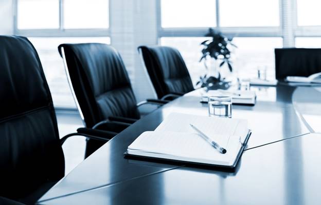 A corporate meeting room setting, with empty leather chairs and a glossy conference table. A notebook and pen sit ready, suggesting a place for important business decisions.